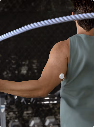 A person using a jump rope during a workout, viewed from behind, wearing a sleeveless top with a small round sensor attached to the upper arm. A row of weights is visible in the background.