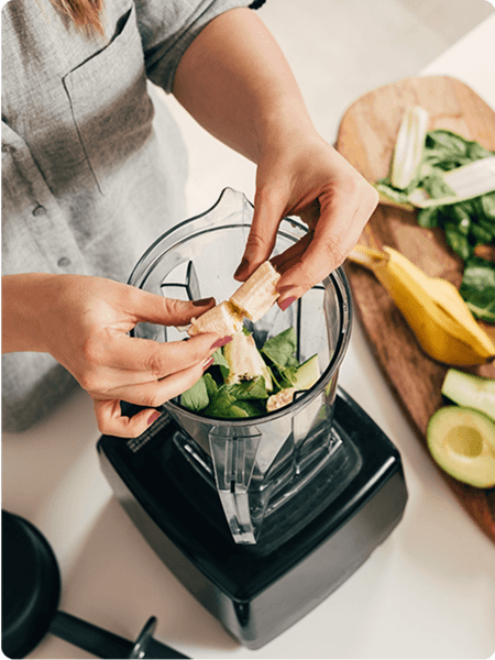 A photo of a person putting a banana into a blender that has other vegetables 