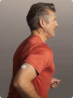 A person running, wearing a orange short‑sleeve shirt, with a Lingo biosensor attached to the upper arm against a clear blue sky.
