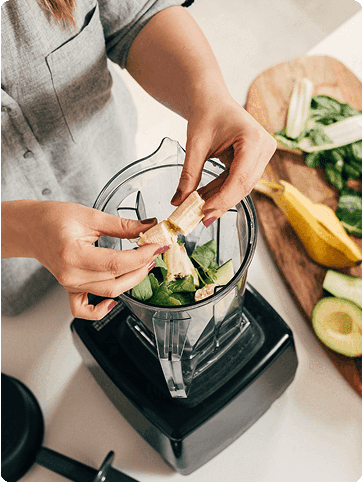 A photo of a person putting a banana into a blender that has other vegetables