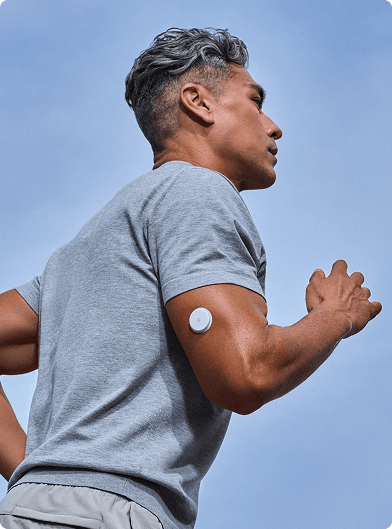 A person running outdoors, wearing a gray short‑sleeve shirt, with a Lingo biosensor attached to the upper arm against a clear blue sky.