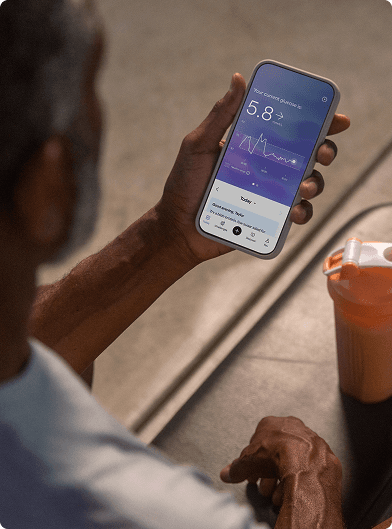 A person holding a smartphone displaying the Lingo app with a glucose reading, seated at a table next to a shaker bottle.