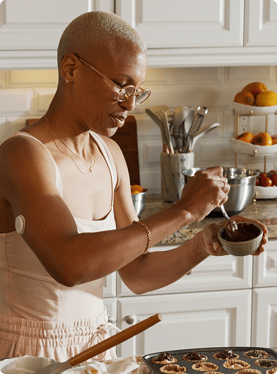 A person in a light-colored kitchen spooning chocolate from a small bowl onto pastries in a muffin tin, wearing a sleeveless top and a Lingo biosensor on the upper arm, with bowls, utensils, and fruit on the counter in the background.