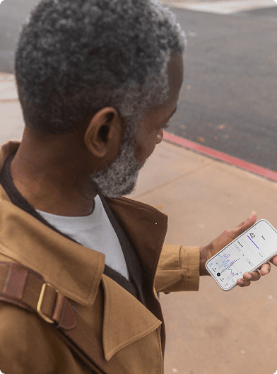 A person standing outdoors on a sidewalk holding a smartphone that displays the Lingo app, wearing a tan coat with a bag strap over one shoulder, with a street curb visible in the background.